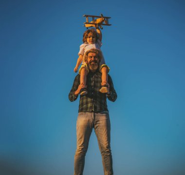 Baba oğul tahta uçakla oynuyorlar. Çocuk babasının omuzlarında oturuyor..