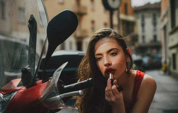 Makijaż w ruchu. Moda dziewczyna z czerwonymi ustami umieścić szminkę patrząc w lustrze motocykla. Moda uliczna. Życie miasta. — Zdjęcie stockowe