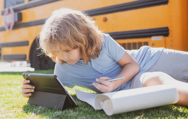 Kind erledigt Hausaufgaben auf Rasen im Park in der Nähe des Schulbusses. Schulkind im Freien. — Stockfoto