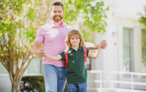 Parent et élève de l'école primaire vont de pair. Professeur en t-shirt et mignon écolier avec sac à dos près du parc de l'école. Un parent mène un petit écolier en première année. — Photo