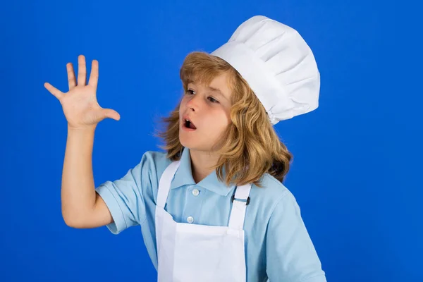 Çocuk aşçı, izole edilmiş mavi stüdyoda yemek hazırlıyor. Çocuklar yemek yapıyor. Önlüklü ve şapkalı genç çocuk mutfakta sağlıklı bir sebze yemeği hazırlıyor.. — Stok fotoğraf