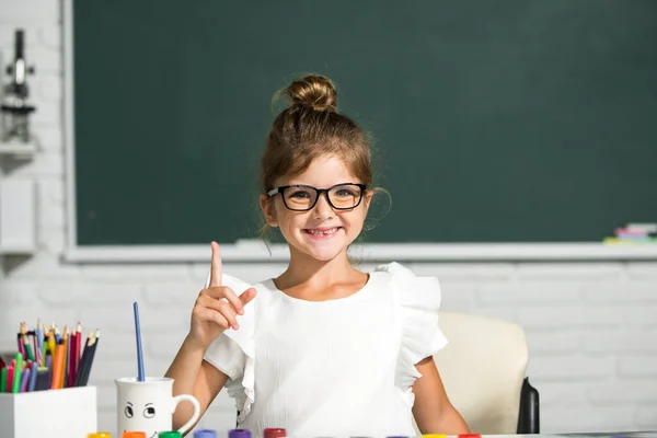 Carino bambino bambina pittura con vernici colore e pennello. Lezione di disegno a scuola. Divertente scuola ragazza faccia con gli occhiali. — Foto Stock