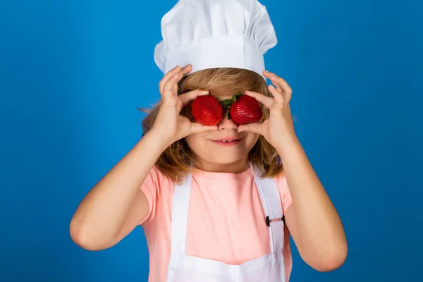 Niño chef aislado en azul. Divertido cocinero niño pequeño cocinero con gorra de cocinero uniforme y delantal mantenga fresas cocinadas alimentos en la cocina. —  Fotos de Stock