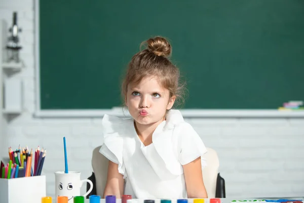 Çocuk kız sınıfta resim çiziyor, masada oturuyor, okulun karatahta arka planında eğleniyor. Komik okul kızı suratı.. — Stok fotoğraf