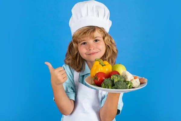 Детский повар держит тарелку с овощами, изолированными на голубом. Забавный маленький повар, одетый в униформу и фартук, приготовленный на кухне.. — стоковое фото