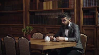 Kütüphanede olgun bir adam ve okumayı seviyor. Takım elbiseli sakallı adam memnun görünüyor. Rahatlama ve rahatlama, hobi konsepti. Edebiyat ve başarı. Kahve ya da çay iç. Kütüphanedeki kendinden emin yetişkin öğrenci