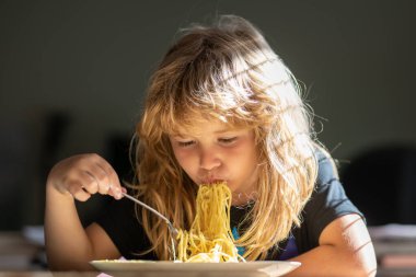 Küçük çocuk mutfakta spagetti yiyor. Masada oturan genç bir çocuk yüzünde komik bir ifadeyle sağlıklı yiyecekler yiyor. Makarna yiyen komik bir çocuğun portresi.