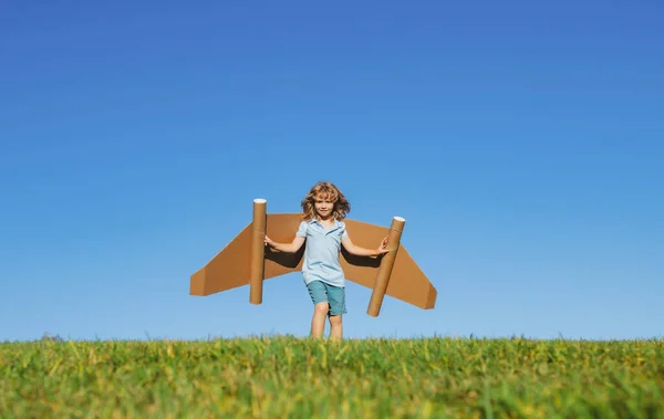 Boy child with wings at sky imagines a aviator pilot and dreams of flying. Kid boy plays pilot with cardboard toy airplane in the park