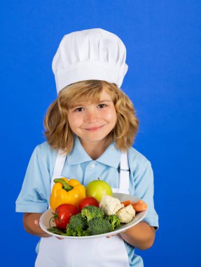 Aşçı şapkalı ve önlüklü çocuk yemek hazırlayan sebzeli yemek tabağı. Mutfakta sebzeli küçük bir aşçı. Doğal çocuk yemekleri.