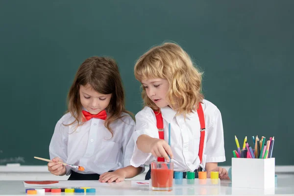 Funny pupils draws in classroom on school blackboard background. School friends kids boy and girl painting together in class. Little artists painting, kids drawing art