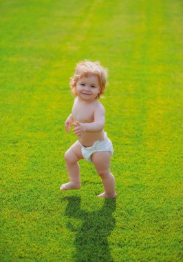 İlk adım. Bebek bez pantolonuyla yeşil çimlerde çıplak ayakla duruyor. Güzel, mutlu küçük çocuk yaz parkında yeşil bir çayırda yürüyor.