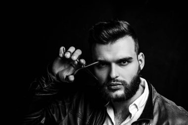 Bearded man, bearded male. Portrait of stylish man with beard. Barber scissors and straight razor, barbershop. Handsome barber is holding scissors near eye on black background. Stylish hairdresser