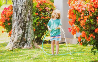 Bir yaz akşamı evin arka bahçesindeki su hortumuyla çiçekleri sulayan sevimli bir çocuk.
