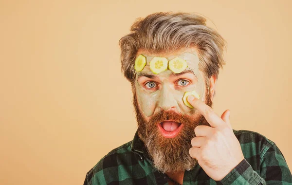 Funny male model with clay mask and cucumber slices on face. Spa, dermatology, wellness and facial treatment concept. Man having cosmetic moisturizing mask, isolated studio background. Male face care