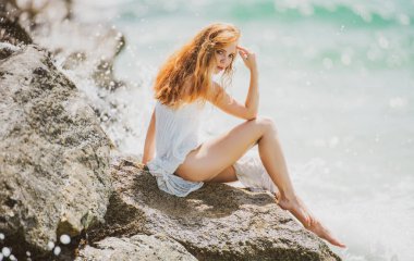 Mavi dalgalara karşı kumlu kayalıklarda oturan sakin kadının yan görüntüsü. Çekici bir genç kadın sahilde seksi bir yaz elbisesi giyiyor.