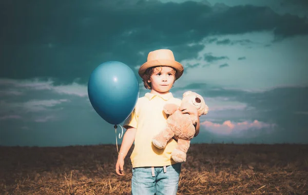 日没の背景におもちゃのテディと風船でフィールドに小さな男の子 庭の外で幸せな男の子 夏の野外で幸せな子供 — ストック写真