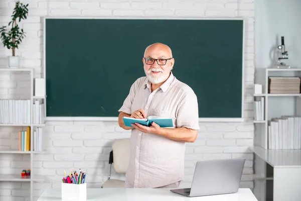 Man mature teacher. Portrait of smart senior tutor in glasses with book on the blackboard in class at high school or college. Aged male retired teacher. Old student study in university