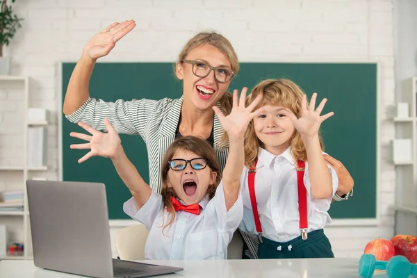 Excited teacher with a school kids learning at laptop computer, studying with online education. Teacher and little students in the classroom. Teacher helping child pupil from elementary school