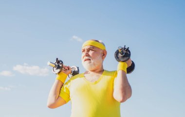 Antrenmandan sonra yaşlı bir adam. Sağlıklı yaşam tarzı neşeli. Kıdemli adam mavi gökyüzünde egzersiz yapıyor. Özgürlük emekliliği kavramı. Elinde dambılla kameraya bakan mutlu son sınıf öğrencisi.