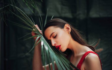 Palmiye yaprağı tutan güzel bir kadın. Güzellik yazı makyaj fotoğrafı. Doğal kozmetik ve cilt bakımı. Harika bir teni ve uzun koyu renk saçları olan güzel bir genç kadın. Palmiye yaprağı