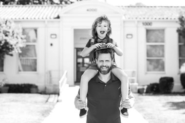 Erkek ve heyecanlı çocuk okula yakın bir yerde sırtına biniyor. Baba oğlunu destekliyor ve motive ediyor. Çocuk ilkokula gidiyor.