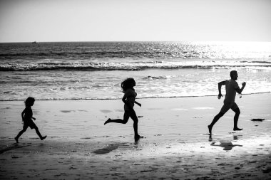 Mutlu genç aile gün batımında sahilde koşup zıplıyor. Aktif ebeveynler ve insanlar çocuklarla tropikal yaz tatillerinde dışarıda etkinlik yapıyorlar.