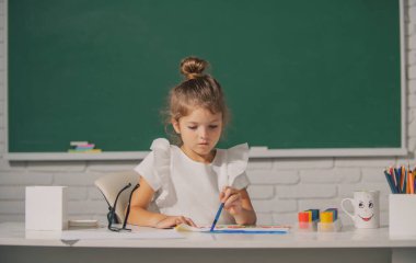 Çocuk kız sınıfta resim çiziyor, masada oturuyor, okulun karatahta arka planında eğleniyor. Komik çocuk suratı
