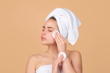 Genç kadın mikiler suyu ve daire şeklinde beyaz ped kullanıyor, spa prosedürlerini seviyor, kafaya havlu sarıp, bej renkli stüdyo arka planında izole edilmiş. Sabah, hijyen, güzellik.