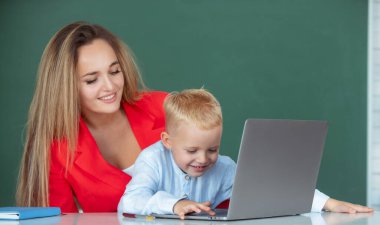 Anne ve oğlu birlikte bilgisayar laptopu kullanıyorlar. Öğretmen okul çocuğuna dersi açıklıyor. İlkokul çocukları öğreniyor.