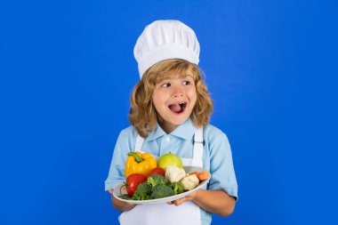 Aşçı çocuk sağlıklı yemek için taze sebze yapıyor. Gri arka planda izole edilmiş küçük çocuk portresi. Aşçı çocuk, sebzeli tabak tut.
