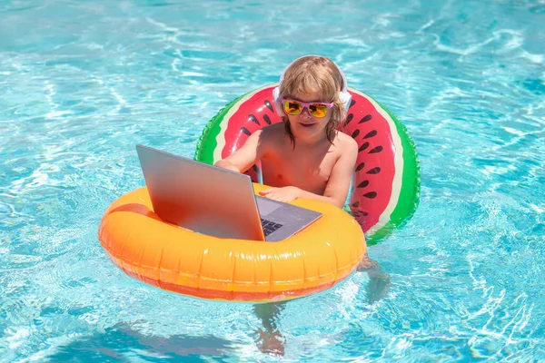 Happy little boy learning online computer laptop. Child relax and rest on colorful inflatable ring using computer in swimming pool. Work outside with laptop in pool