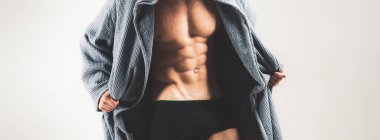 Güçlü atletik adam. Fitness modeli, mükemmel kas gövdesini gri arka planda fotokopi alanı ile izole edilmiş olarak gösteriyor. Kaslı model genç adam bornozunu çıkarıyor.