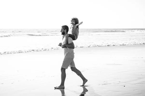 父と息子は海を歩いている 若い男の子の息子を運ぶハンサムな男の父親 幸せなパパが子供を抱いてる 夏の日に子供の男の子とパパ フレンドリーな家族と夏休みの概念 — ストック写真