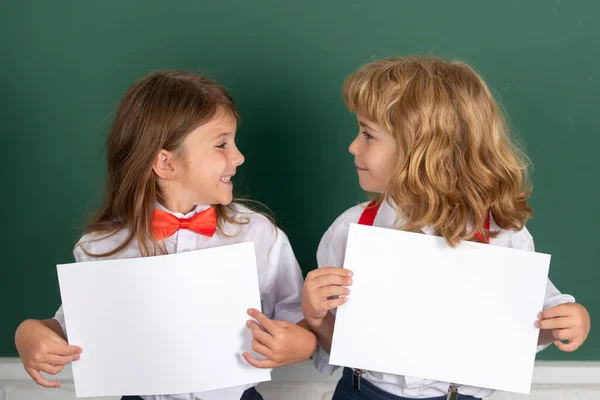 Schoolkids holding white paper blank, poster with copy space. Happy girl and boy school friends. Face portrait of two schoolkids. Children couple at primary school classmates, young students