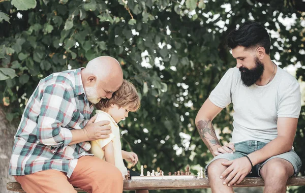 Young boy with father and grandfather enjoying together in park. Old man with little boy playing chess