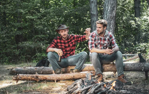 Happy young people camping in woods. Summer lifestyle. Young guy having a picnic. Enjoying camping holiday in countryside. Two Happy people sitting around campfire with beer