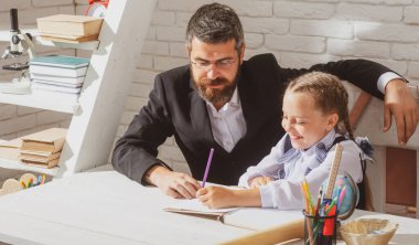Öğrenci kız, öğretmeniyle birlikte ders çalışıyor ve yazı yazıyor. Sevimli öğrenci kız okul malzemeleriyle masasının yanında gülen yüz ifadesiyle ders çalışıyor.