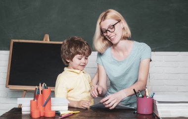 Anne okulda sınıfta oğluna ders veriyor. Komik küçük çocuk ve genç kadın öğretmen kara tahta arka plan üzerinde eğleniyor. Okul çocukları