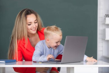 Anne ve oğlu birlikte bilgisayar laptopu kullanıyorlar. Küçük okul çocuğu annesiyle dizüstü bilgisayar kullanıyor. Öğretmenler okulda öğrencilere yardım ediyor.