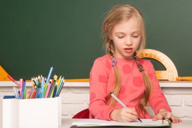 İlkokuldan bir çocuk ilkokulda masada oturuyor, not defterine yazıyor ya da çiziyor. Okulda tahtaya yakın bir okul çocuğu.