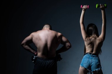 Çıplak vücutlu kaslı bir adam, arka planda koyu renkli halterleri olan fitness kadını. Dambılla egzersiz yapan seksi spor çifti. Zayıf ve sağlıklı seksi kız güçlü kaslı erkek egzersizi yapıyor.