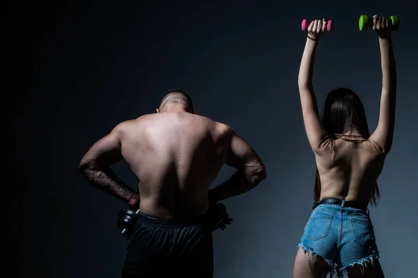Muscular man with naked body, fitness woman with dumbbells on a dark background, back view. Sexy sport couple exercising with dumbbell. Slim and healthy sexy girl with strong muscular man workout