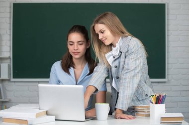 Students girls friends looking at laptop computer in classroom at school college or university on blackboard background. Two students doing homework together and helping each other