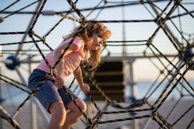 Kid boy climbing the net. Child playing on outdoor playground. Kids play on school or kindergarten yard. Kids play and climb outdoors on sunny summer day
