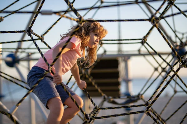 Kid boy climbing the net. Child playing on outdoor playground. Kids play on school or kindergarten yard. Kids play and climb outdoors on sunny summer day