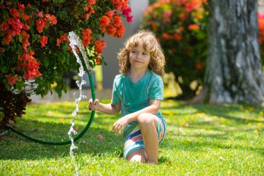 Çocuk bahçede bahçe hortumuyla oynuyor. Komik heyecanlı çocuk arka planda su püskürterek eğleniyor. Yaz çocukları açık hava aktivitesi