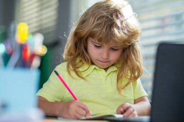 Serious school child writing homework in school class. Portrait of school kid boy siting on table doing homework. Child holding pencil and writing. Boy drawing on white paper at the table