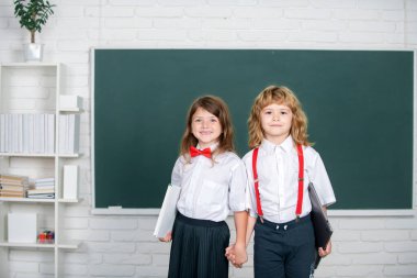 Portrait of two happy school kids studying in classroom at elementary school. Concept of friendship and classmate. Best school friends holding hand near blackboard