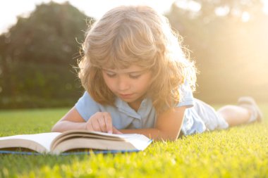 Kid boy reading a book lying on grass. Cute little child in casual clothes reading a book and smiling while lying on grass in park. Smart clever Kids
