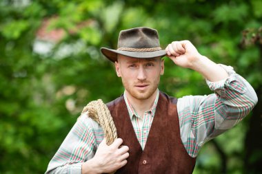 Cowboy farmer man in country side wearing western cowboy hat. Cowboy with lasso rope on green background. American Male model in countryside on farm